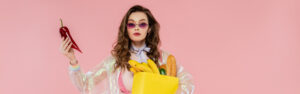 housewife concept, attractive young woman in sunglasses carrying paper bag with groceries and holding red pepper, posing like a doll on pink background, conceptual photography, banner